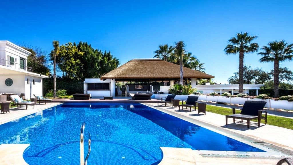 Modern outdoor poolside thatched gazebo with kitchen and seating area.